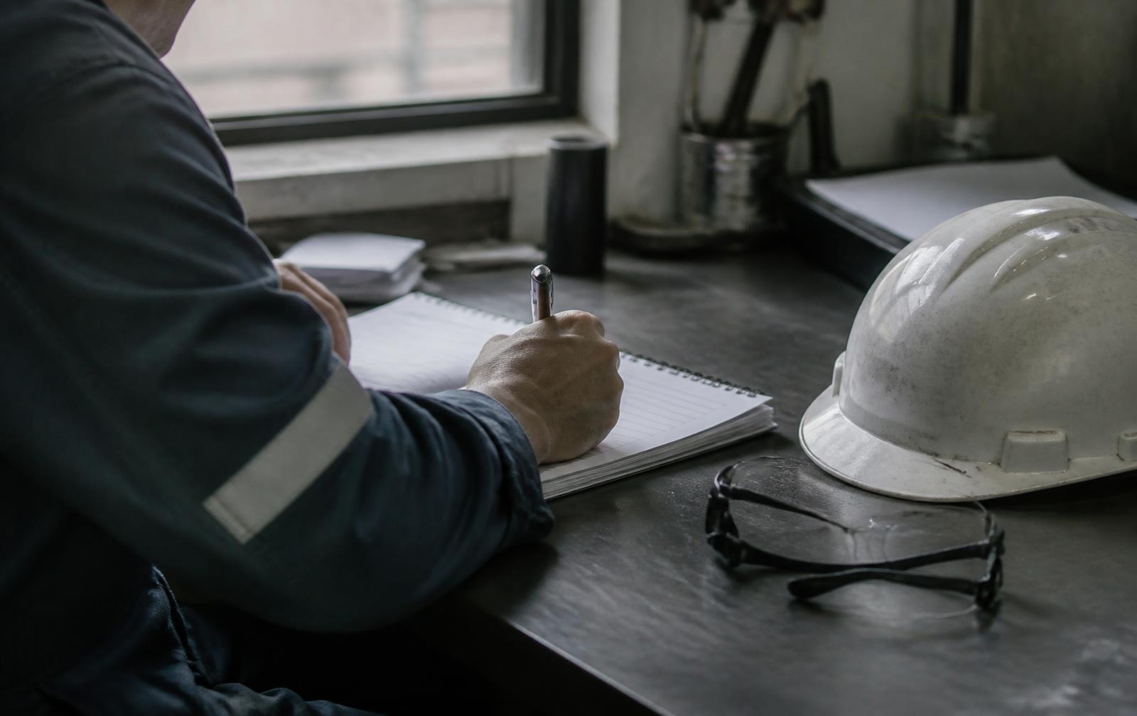 Inspector writing a report at a shop desk, hard hat and safety glasses set down beside the notebook
